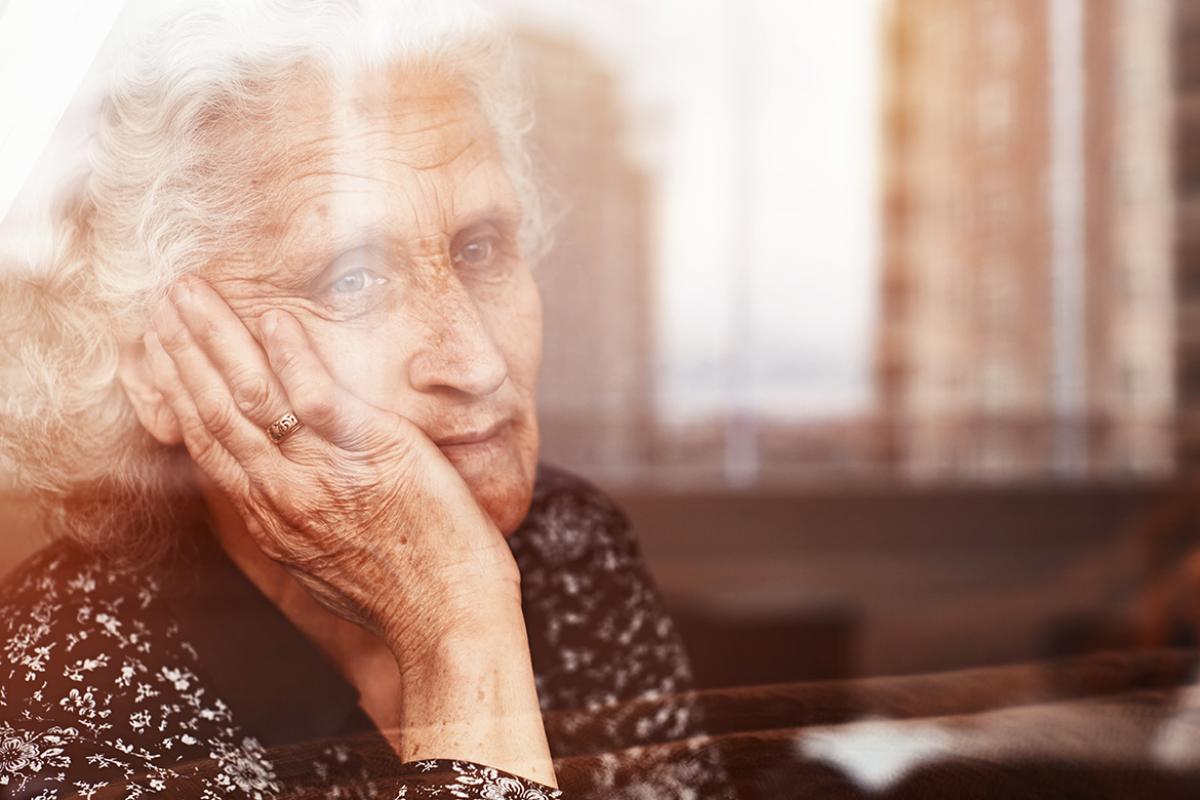 Elderly person sitting down looking out a window with right palm resting on right cheek.