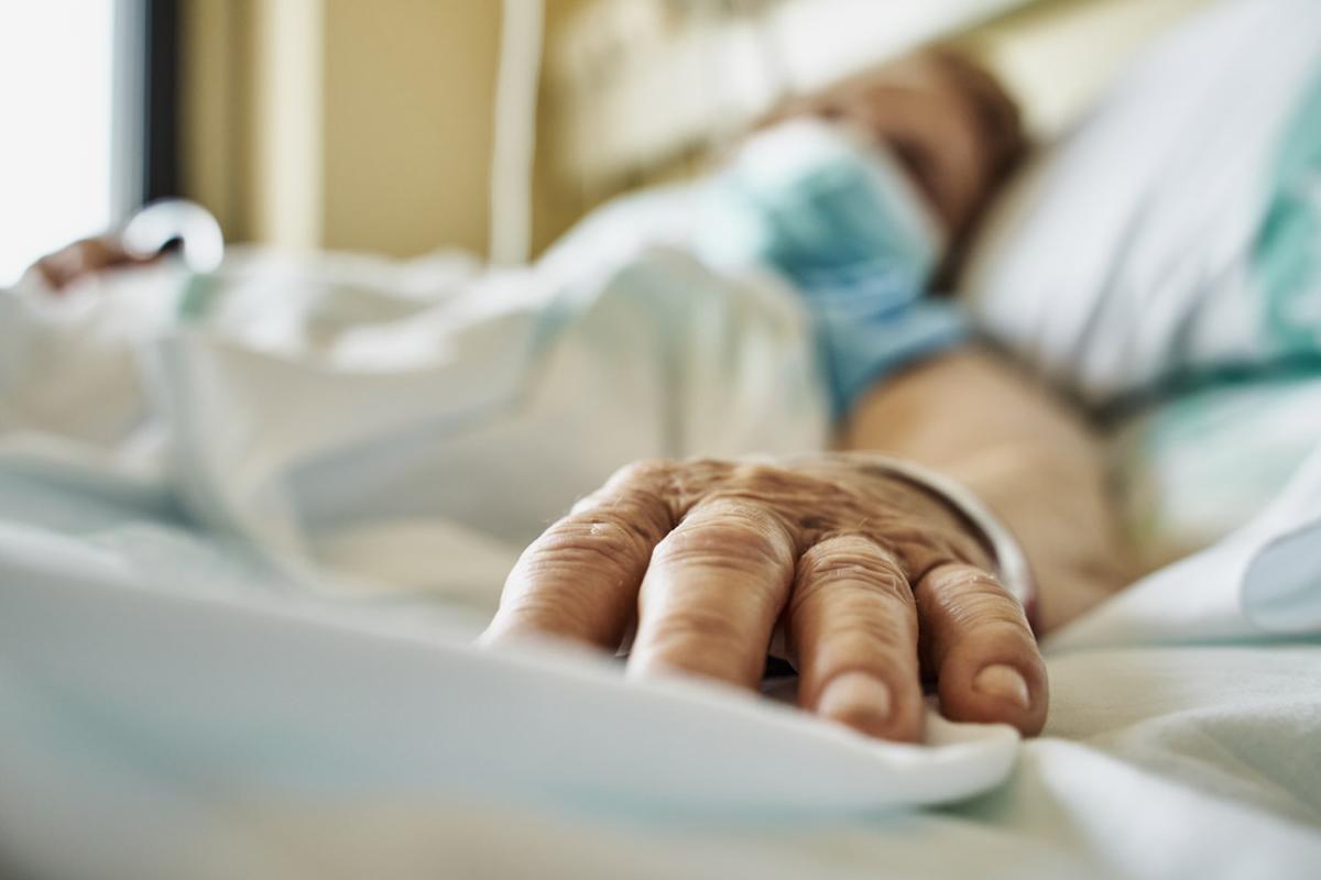 A person wearing a mask laying in a hospital bed, their hand is in focus but the rest of them is blurred.
