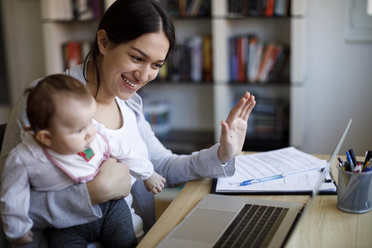 Smiling woman with baby sitting on her lap waving to someone on a laptop computer screen.