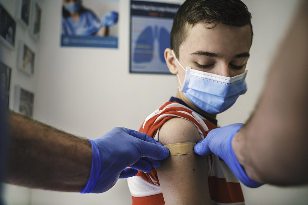 Young boy wearing face mask while a health care worker places a bandaid on his right arm over a vaccination.