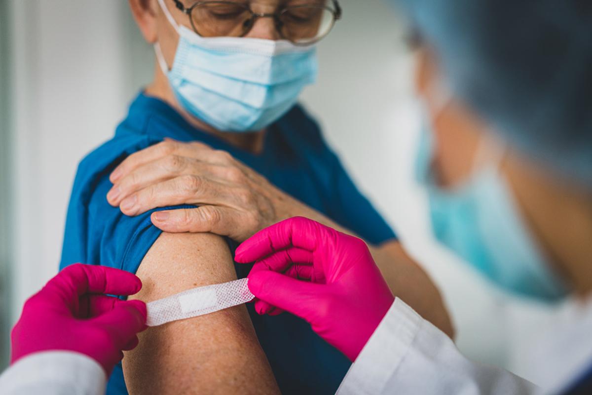 Health professional applying an adhesive bandage to a patient's arm after giving a vaccination, both wearing masks