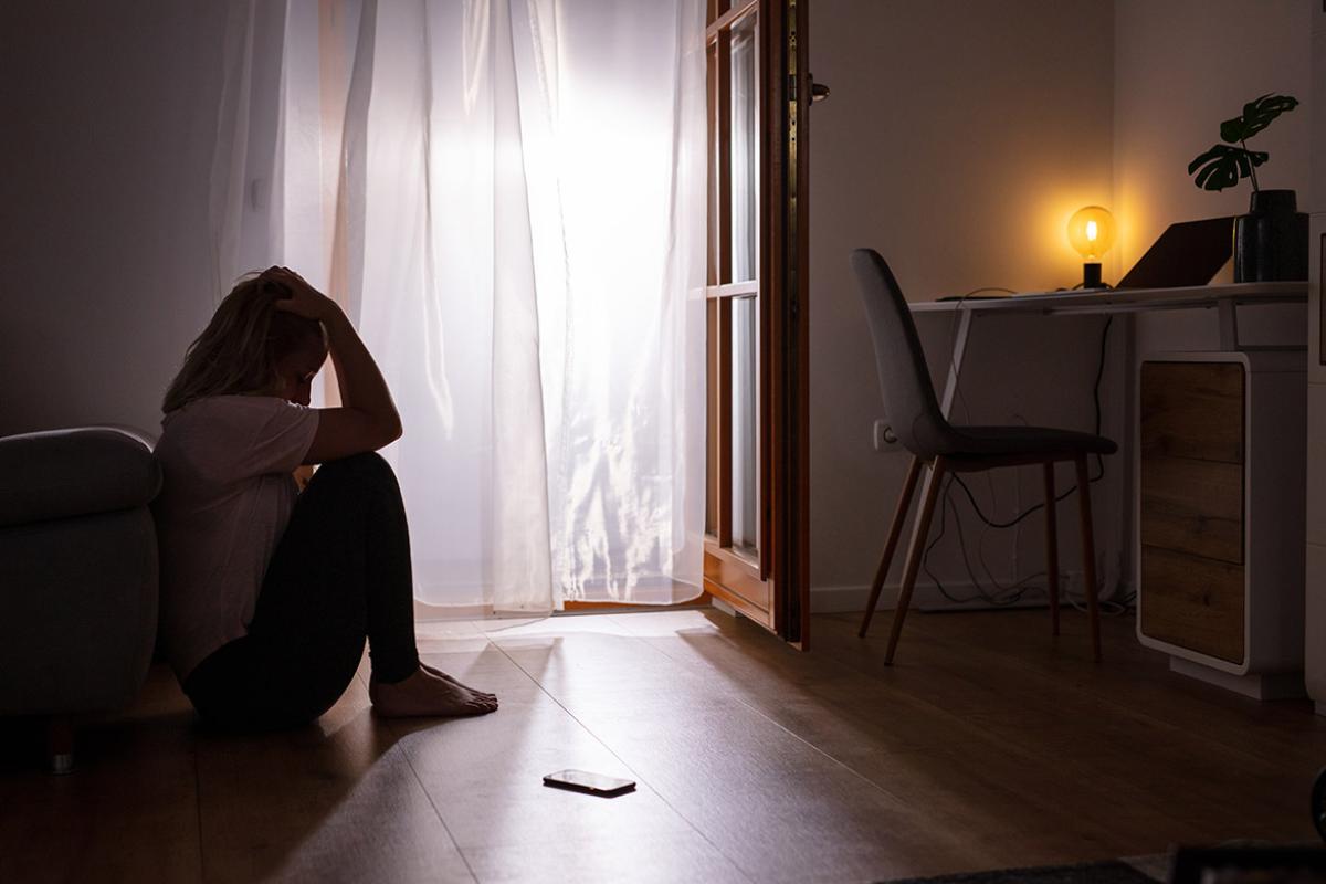 Woman sitting on the floor in the dark with a curtained open patio door, a cell phone to her side