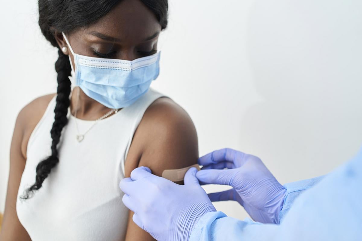 A masked young woman getting an adhesive bandage placed on her left arm after getting a vaccine 