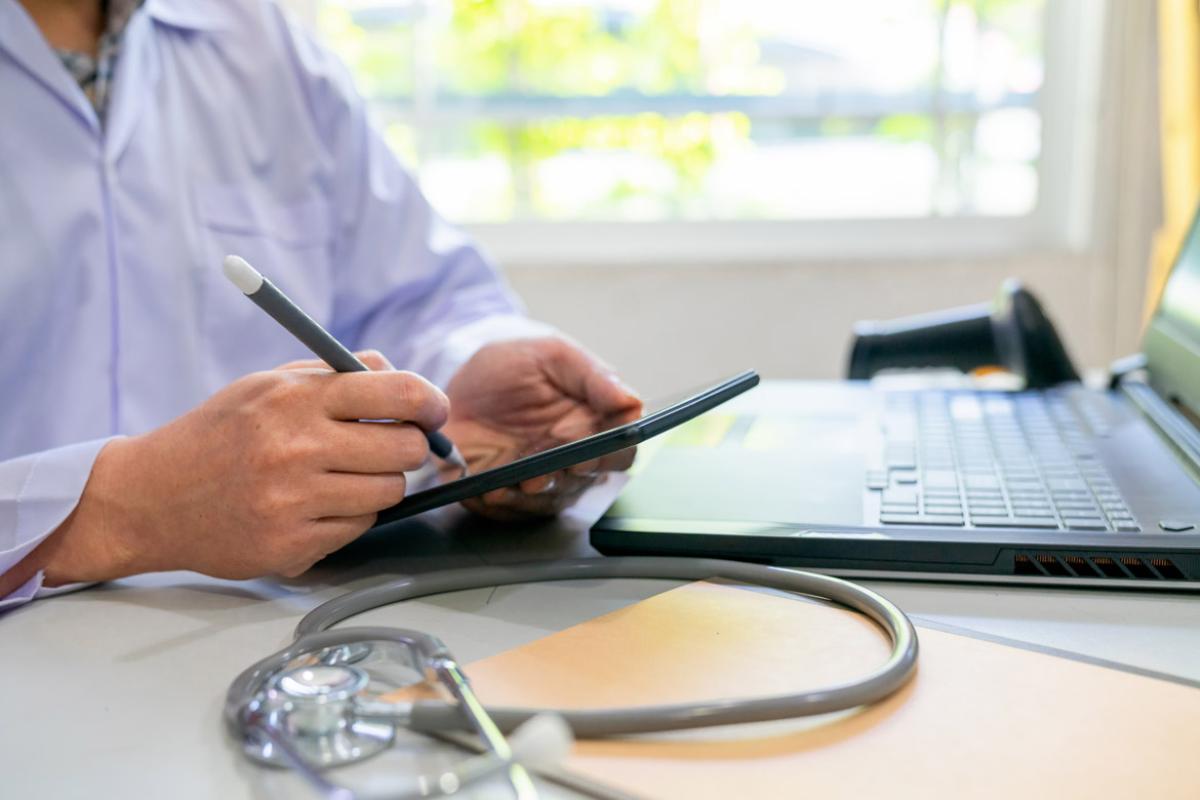 Physician writing on computer pad with stylus pen and an open laptop on top of the desk