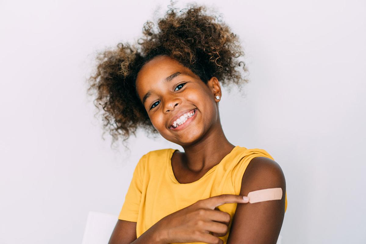 Smiling young girl pointing to an adhesive bandage on her left arm after receiving a vaccination