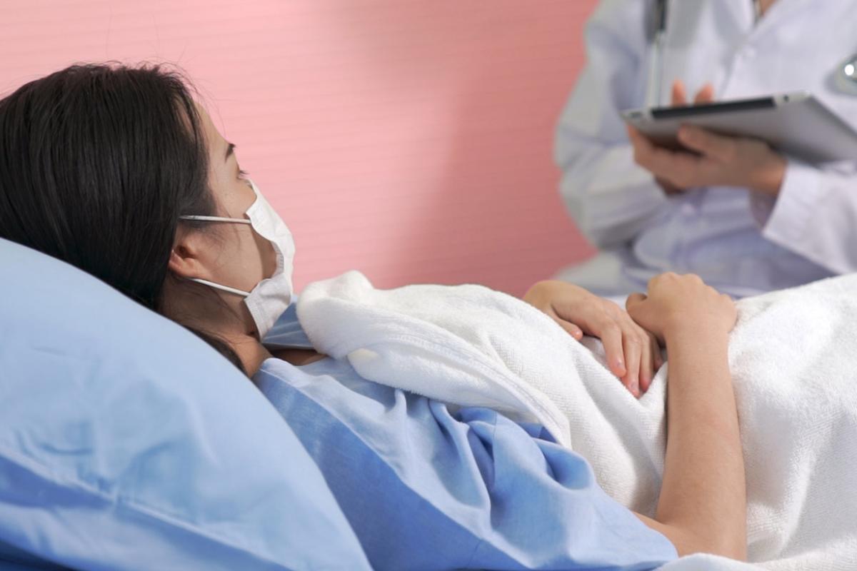 Female patient lying in hospital bed wearing a mask and a physician at her side taking notes on a tablet