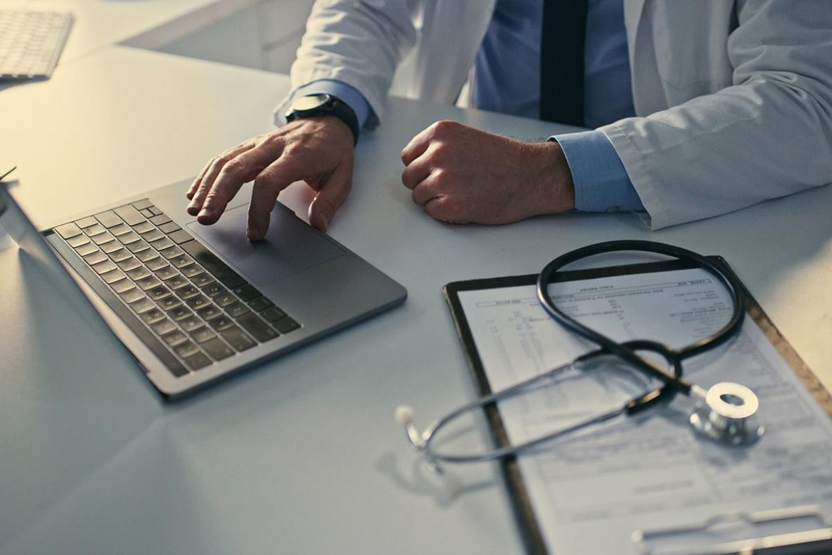 Health care professional working at a laptop with a stethoscope on a clipboard nearby