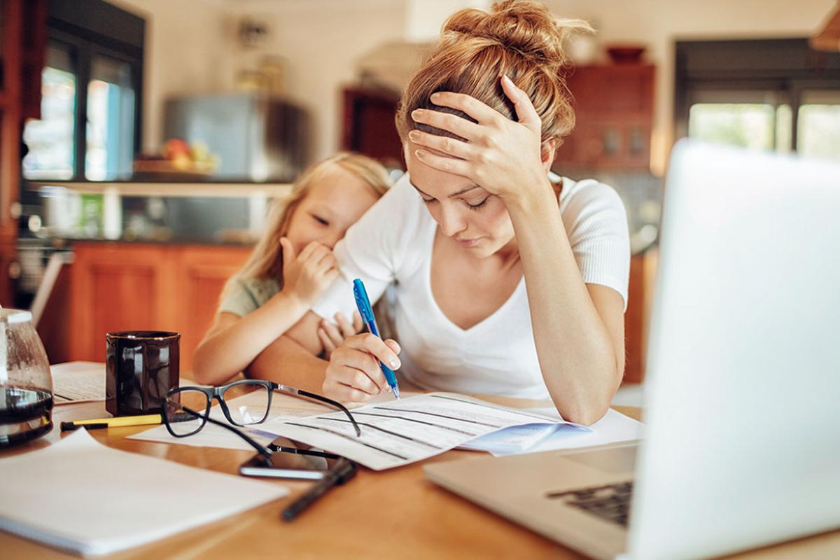Woman sitting at a table with head in hands, child clinging to her arm