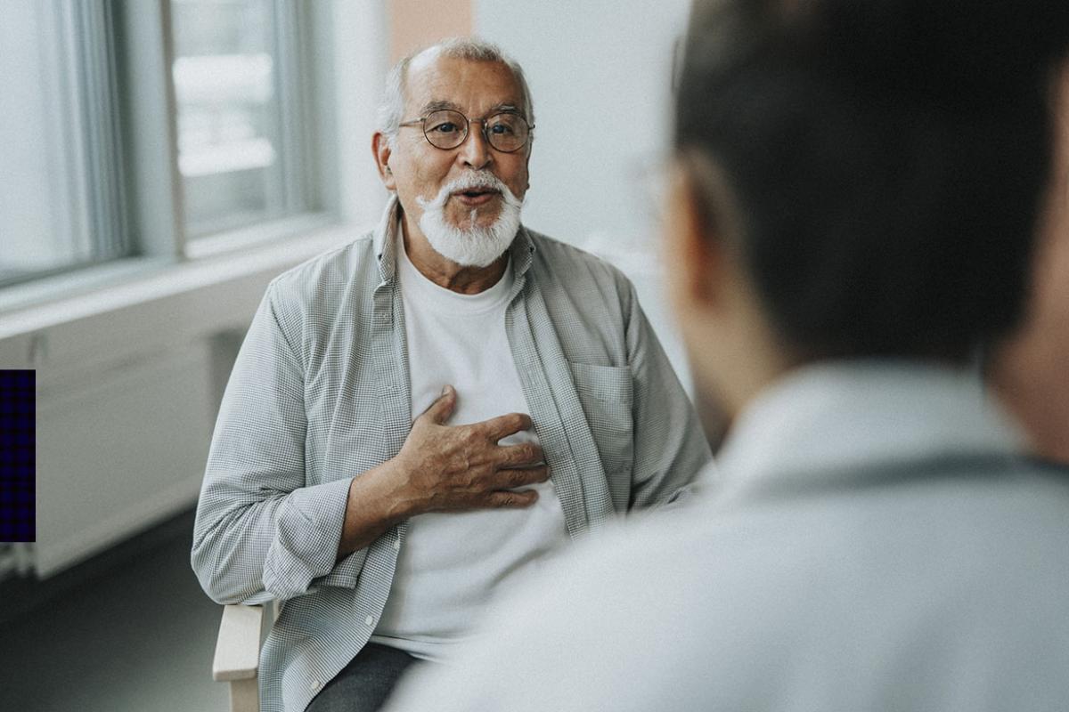 Older man with white hair and beard puts hand on chest while facing doctor