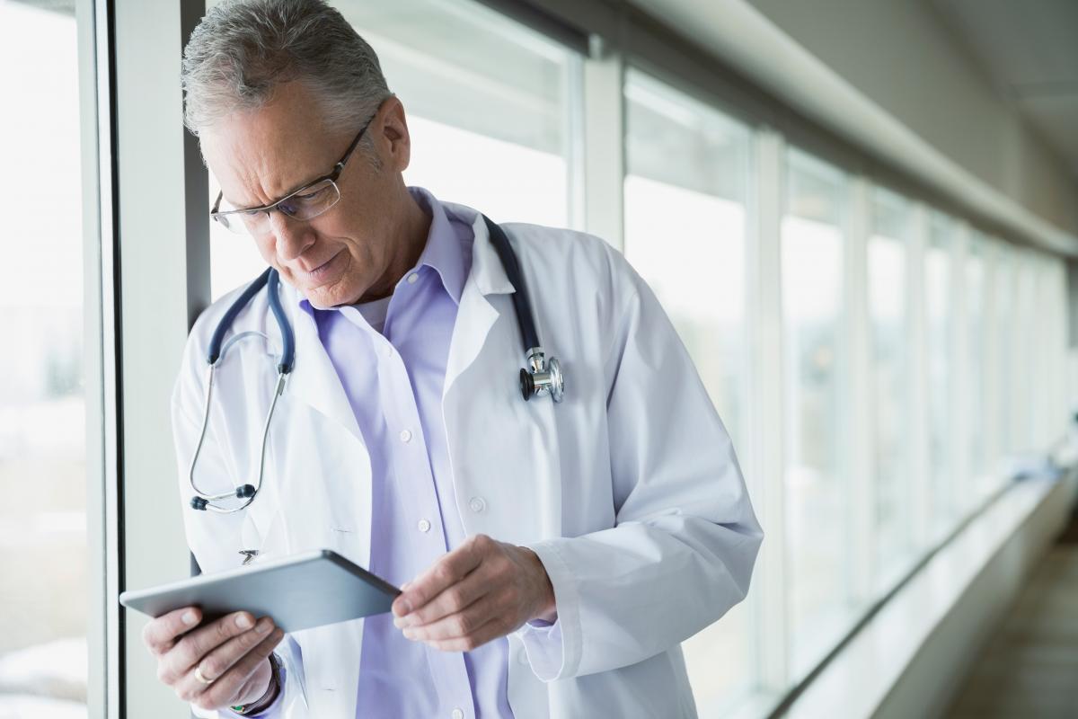 A physician stands in a window-lined hallway and looks at his tablet.