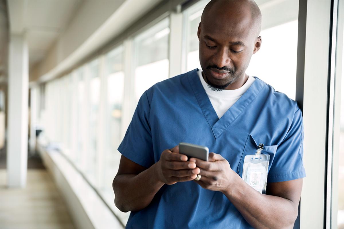 A young physician leans against a wall of windows and looks at his smart phone.