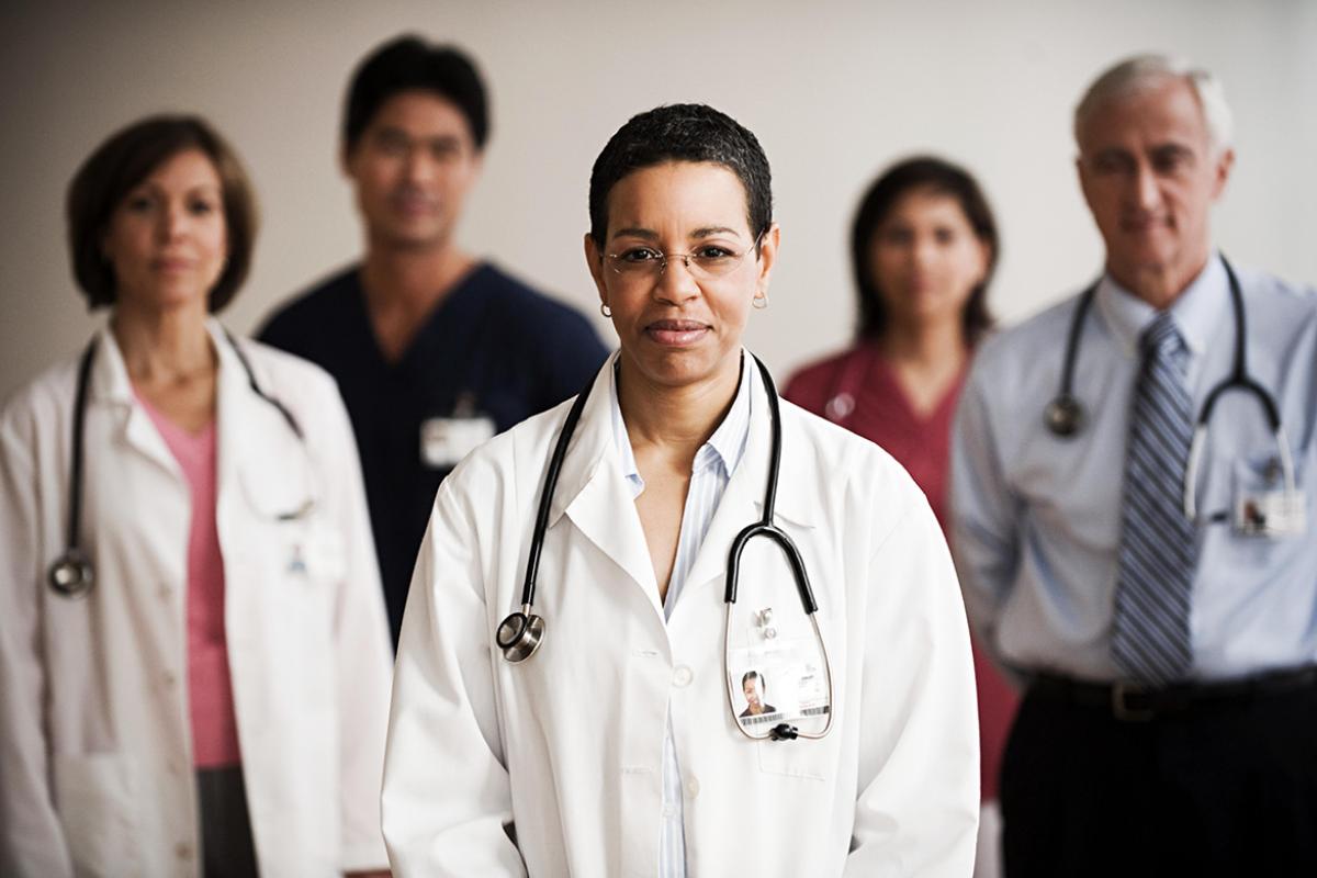 A female physician in a white coat with a stethoscope around her neck faces the camera, with 4 other physicians and medical personnel behind her.