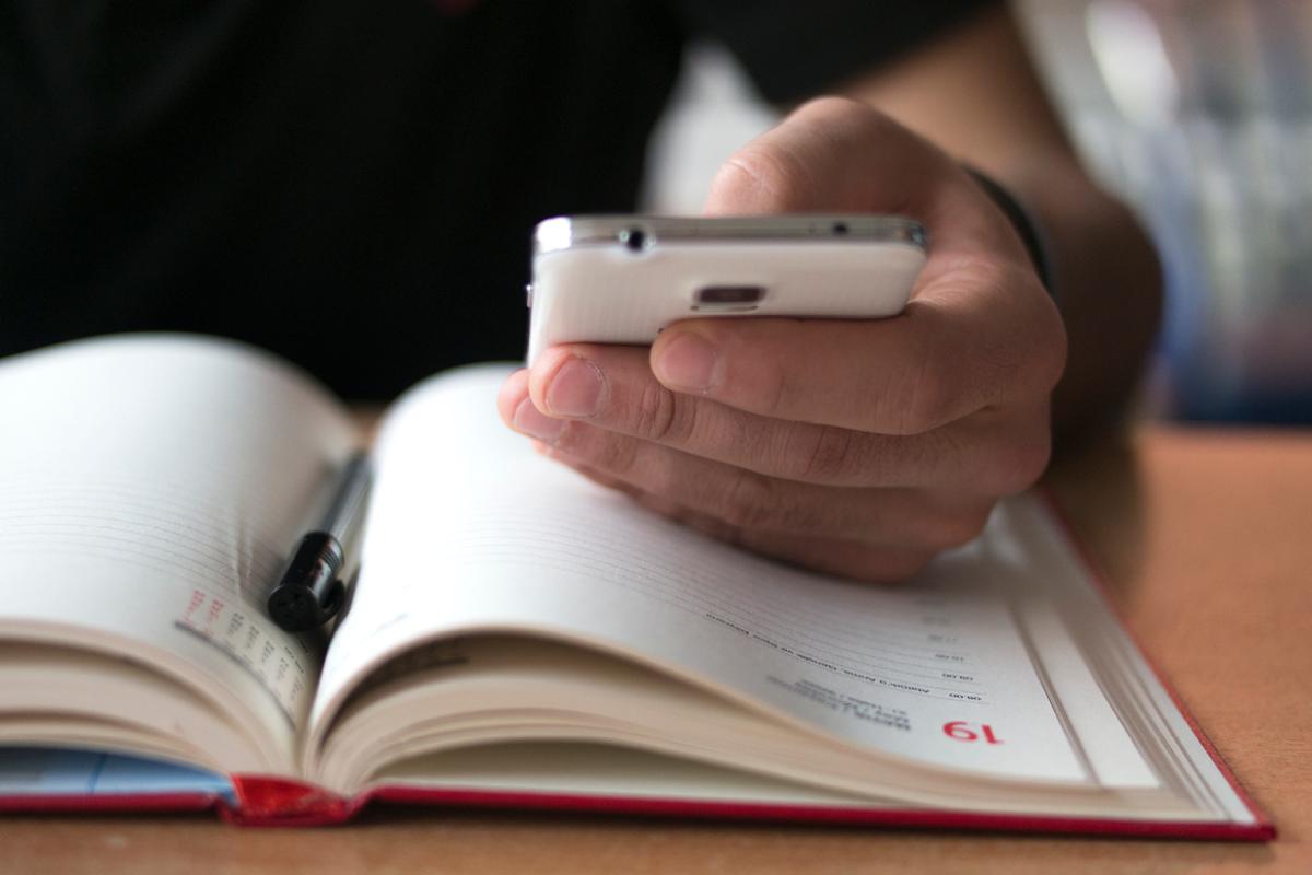 A man looks at his smartphone while making notes in his paper calendar.