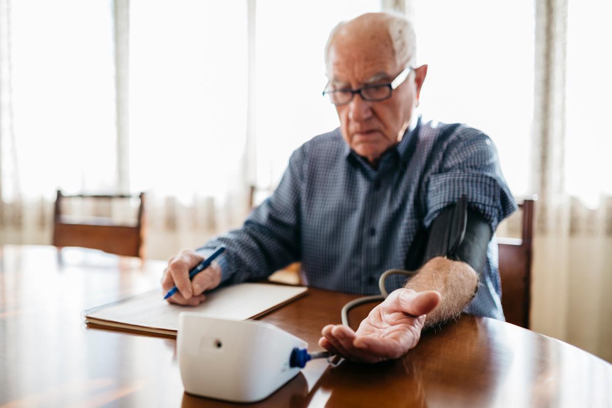 An old man gives himself a blood pressure test and is taking notes on what he sees.