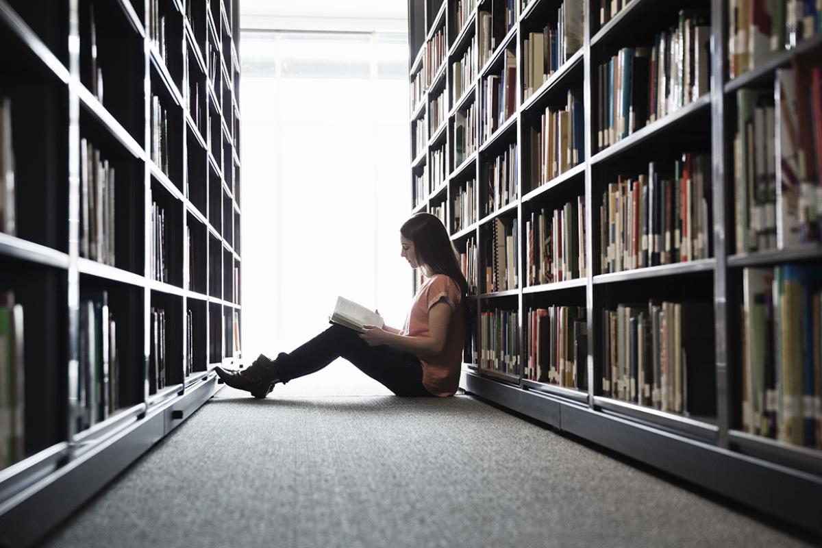 Student reads book between aisles in library.