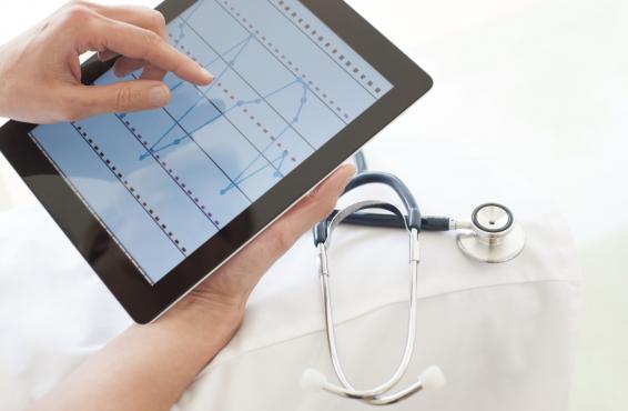 Person holding tablet that shows waves on the screen, next to white coat and stethoscope