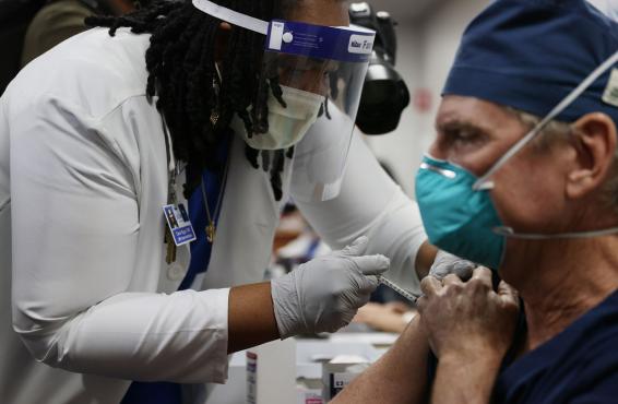 A health care worker in PPE giving a vaccination to a physician in a mask.