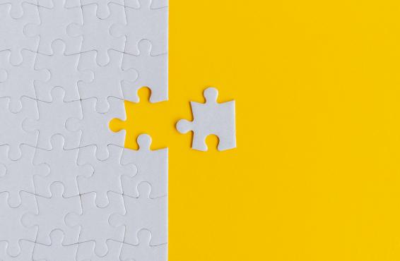 Against a bright yellow background, a close-up of an entirely grey jigsaw puzzle with one piece removed and set to the side of the rest of the puzzle.