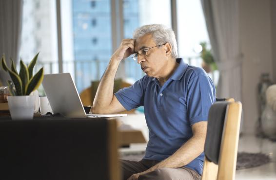 Man in glasses sitting at home at his desk looking at his laptop with his hand on his forehead.