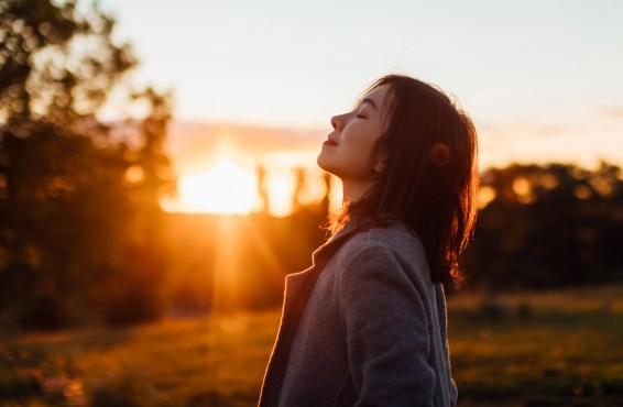 A woman standing in a field, smiling and inhaling the air, as the sun sets behind her.