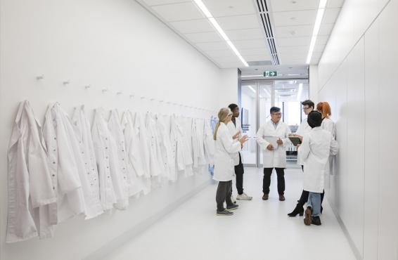 Group of physicians standing in a corridor with white physician coats hanging on racks on a left side wall.