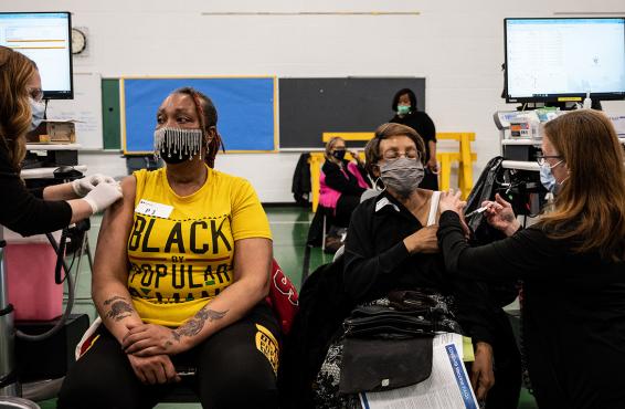Two women wearing masks and receiving vaccinations at a school or communinity center.