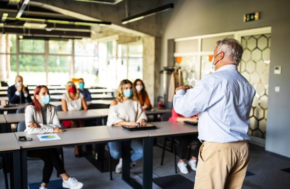 Man making a presentation in front of a group of college age people, all wearing masks