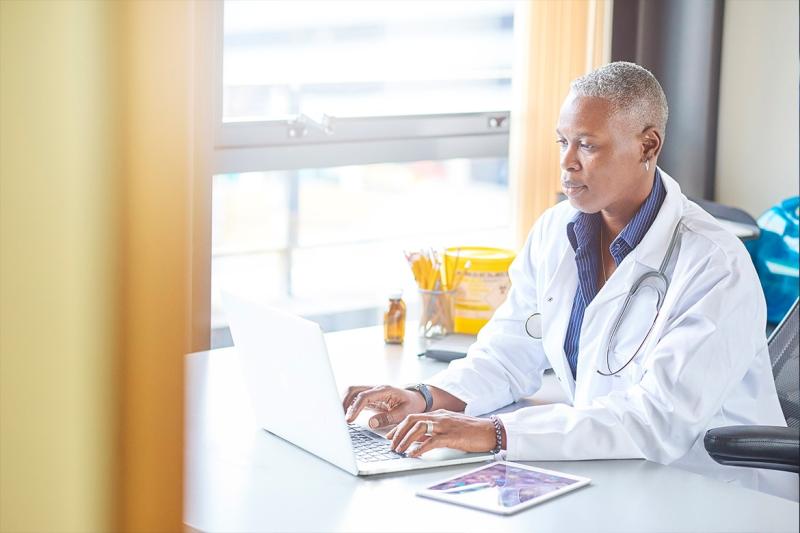 Physician sitting at desk working on laptop with iPad on the side.
