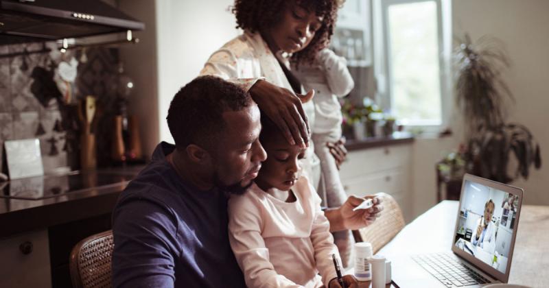 Family sitting around a laptop at a telehealth meeting with a doctor on the laptop screen